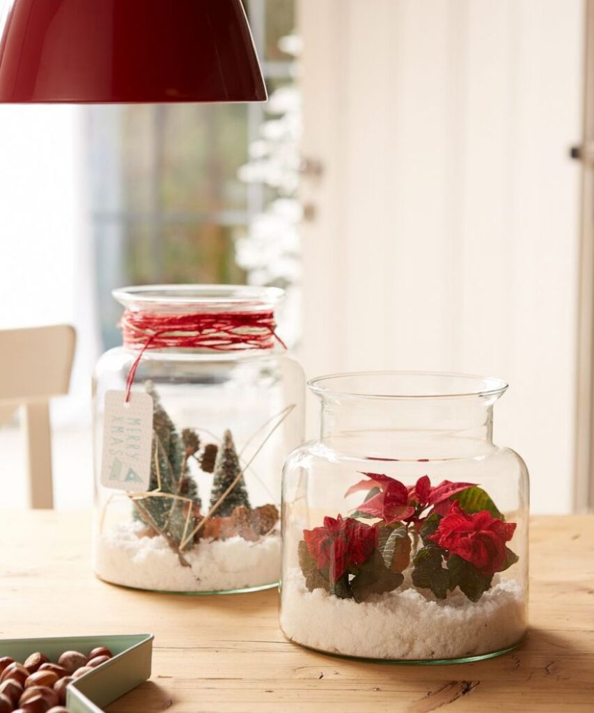 Два стъклени буркана, пълни с коледни украшения. Единият буркан съдържа червени мини коледни звезди и изкуствен сняг, а другият – мини елхички, които придават зимна атмосфера на масата.  