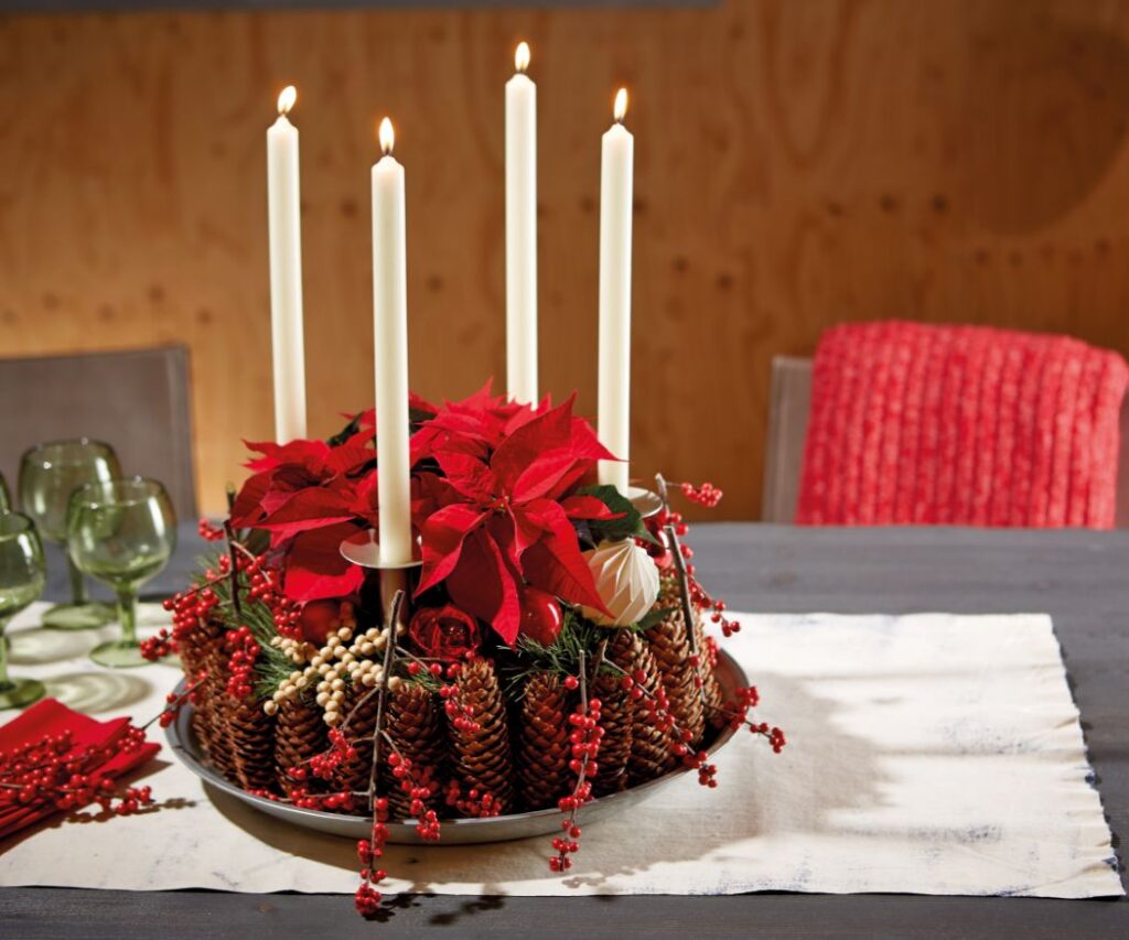 Адвентен венец от червени мини коледни звезди и бели свещи, поставен на маса. Комбинацията от свещи и коледни звезди създава топла, празнична атмосфера.