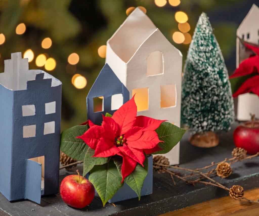 Близък план на селцето от рециклирани материали показва боядисани картонени къщички, мини поинсетии и естествени декорации, които създават коледна атмосфера. 