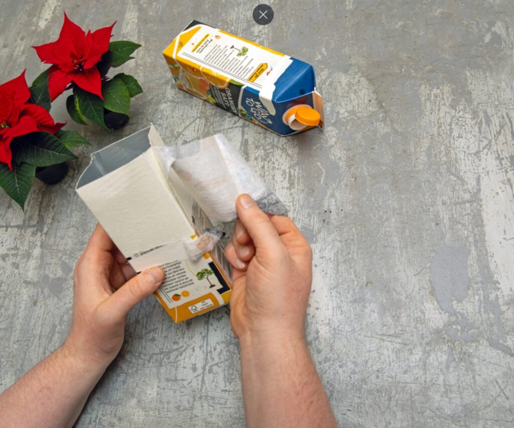 Първата стъпка в този коледен проект за ъпсайклинг, който може да се прави с деца. Двойка ръце премахва външния слой на картонена опаковка за напитка. 