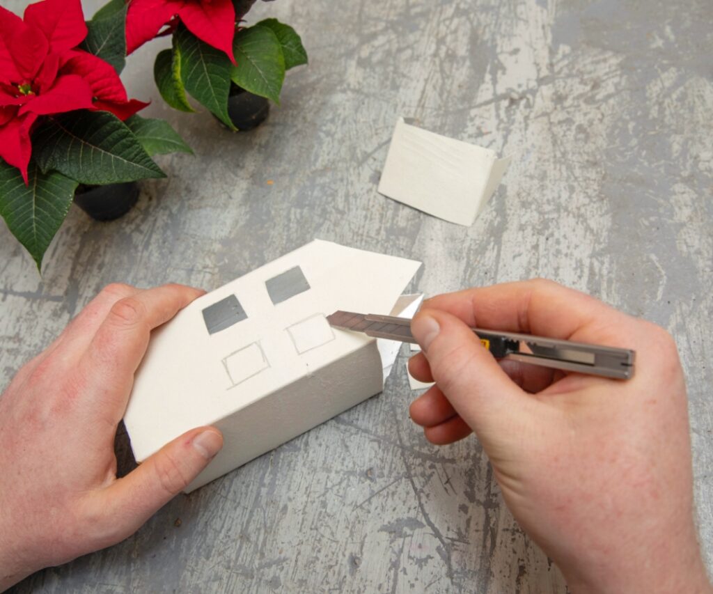 Човек използва макетен нож, за да изреже малки прозорци в картонена опаковка за напитки като част от творческа идея за ръчно изработена къщичка. 