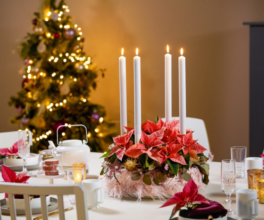 A white-clothed table with an Advent wreath adorned with blush-coloured poinsettias and four lit candles, with a decorated Christmas tree in the background.