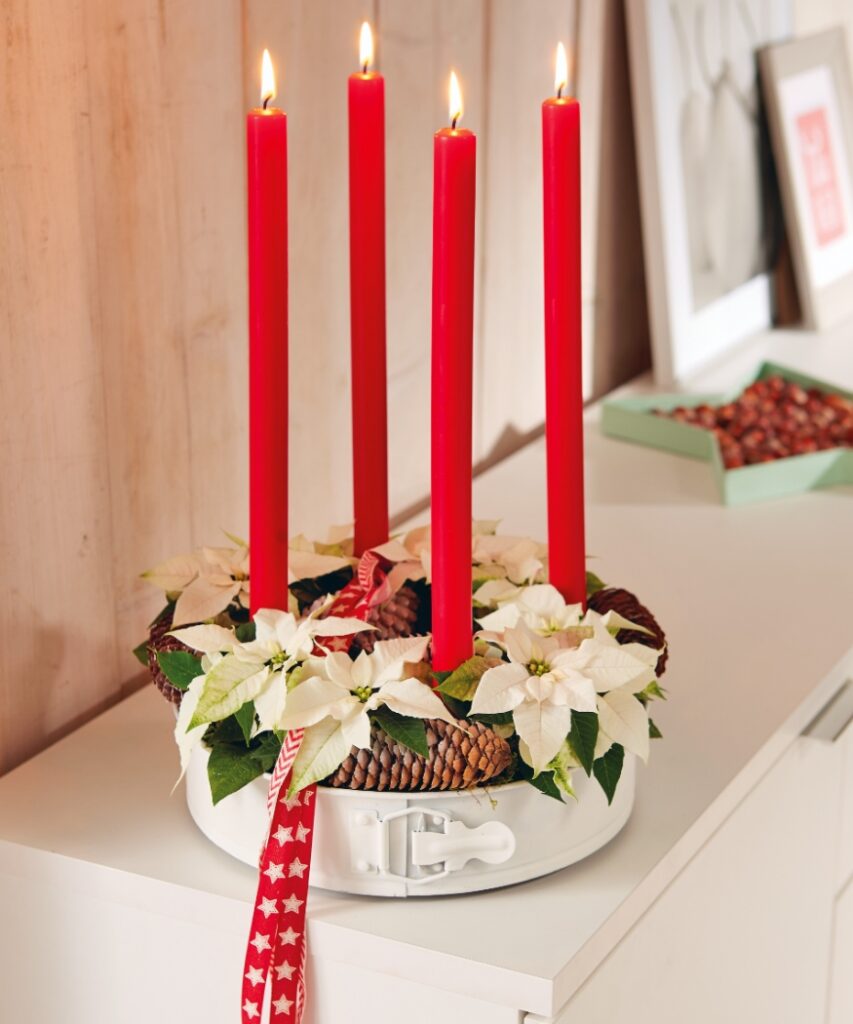 An upcycled cake tin Advent wreath, featuring red candles, white mini poinsettias, moss and pine cones.