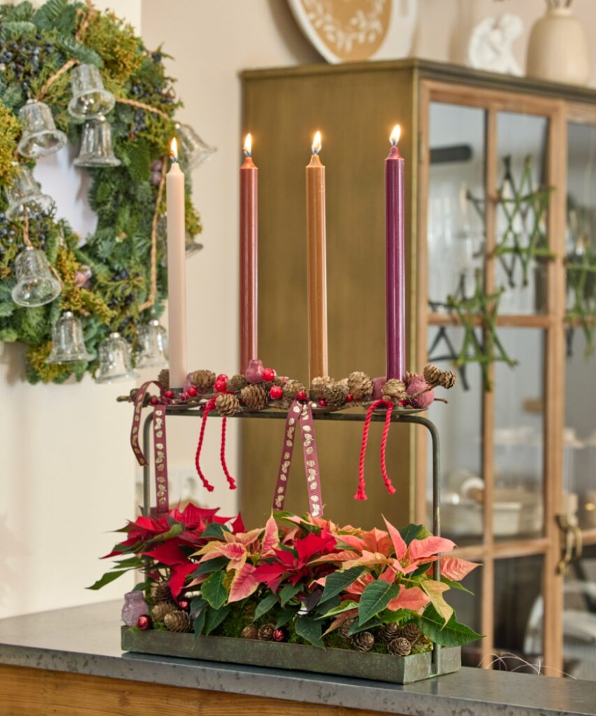 A slim Advent decoration featuring four candles on a metal stand above a planter filled with red and pink mini poinsettias.