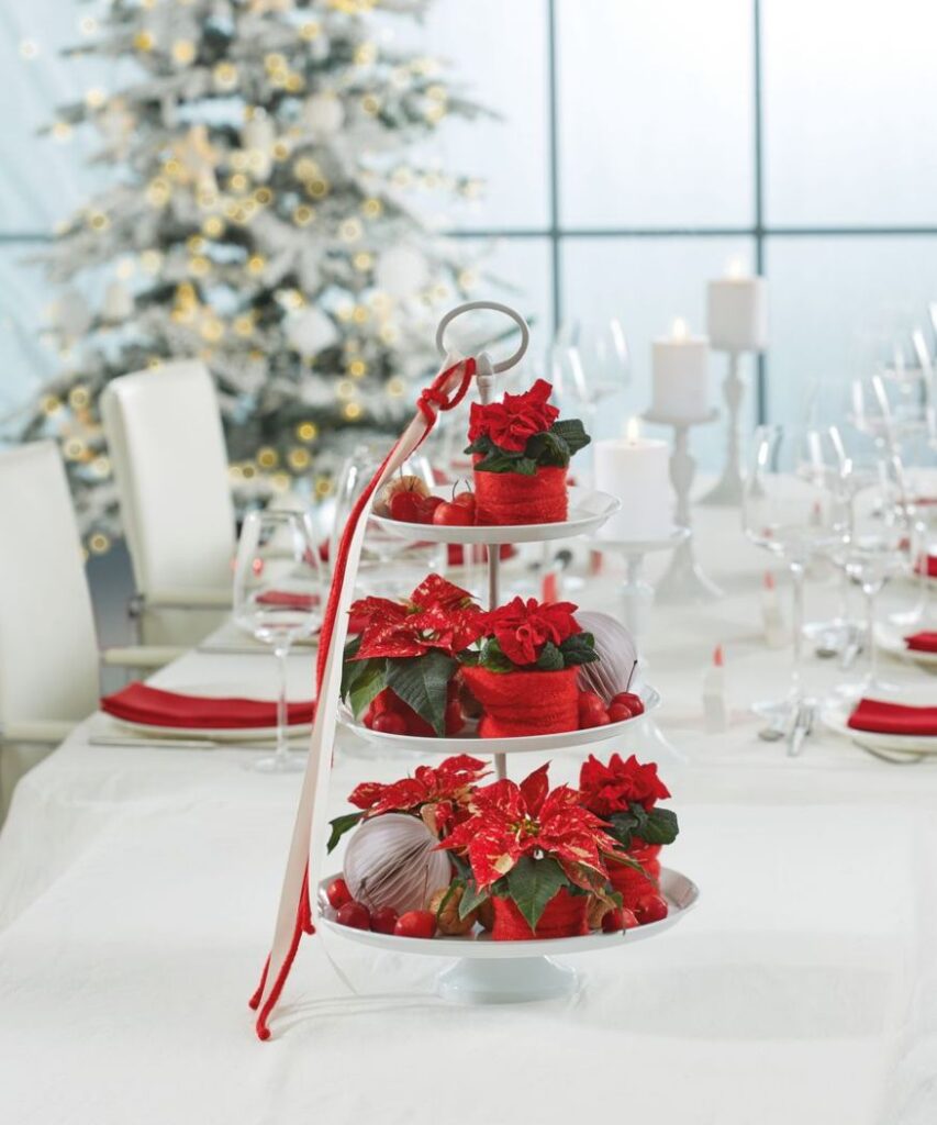 Una decoración de mesa de varios niveles de mini poinsettias rojas delante de un árbol de Navidad decorado.