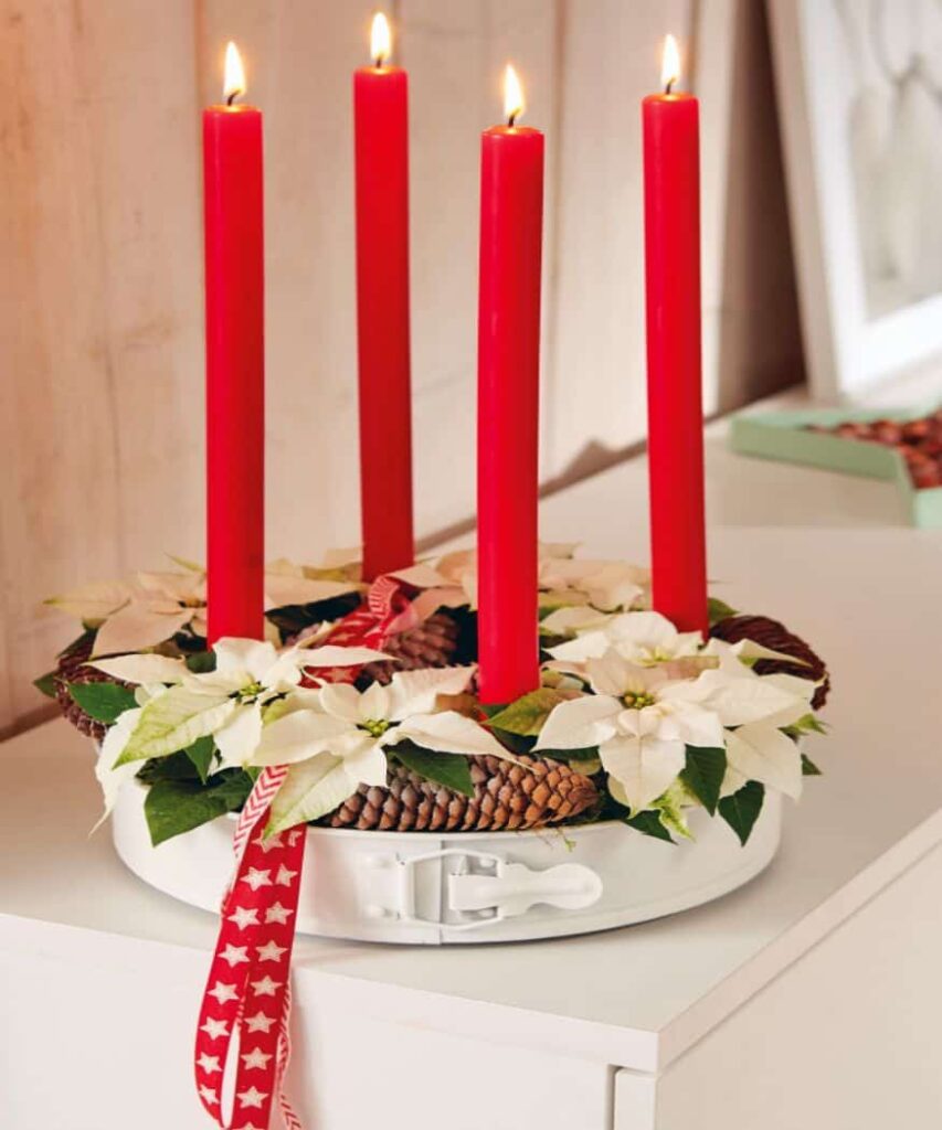 Adventski vijenac od bijelih mini božićnih zvijezda s četiri upaljene crvene svijeće stoji na komodi. Dekorativna crvena vrpca upotpunjuje blagdanski aranžman. Ideja za rukotvorine s kalupom za pečenje.