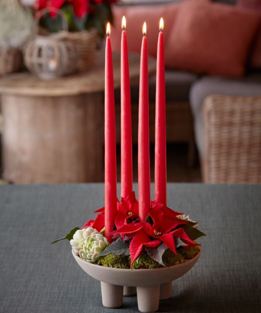Egy adventi koszorú négy skarlátvörös keskeny gyertyával és piros mini mikulásvirággal, egy modern tálban. 