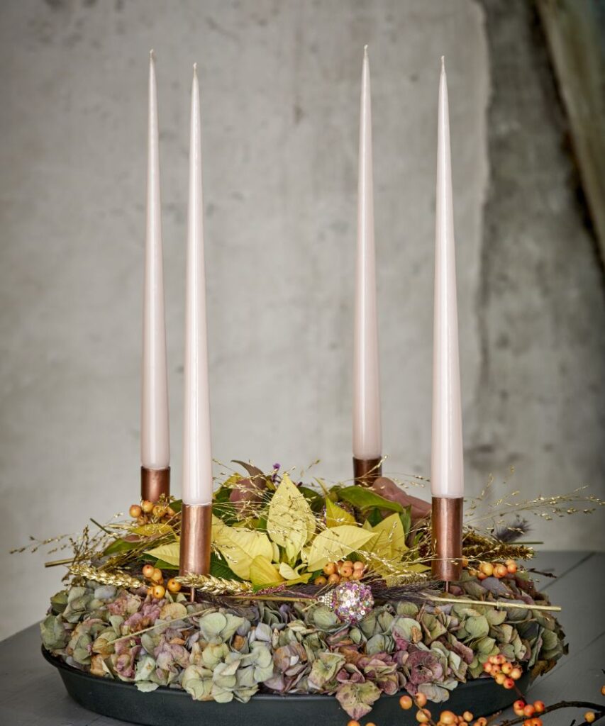 Egy elegáns adventi koszorú, rózsaszínes fehér gyertyákkal és natúr színű, szárított hortenzia virágokkal. A középen egy tejszínű sárga mini mikulásvirág áll. 