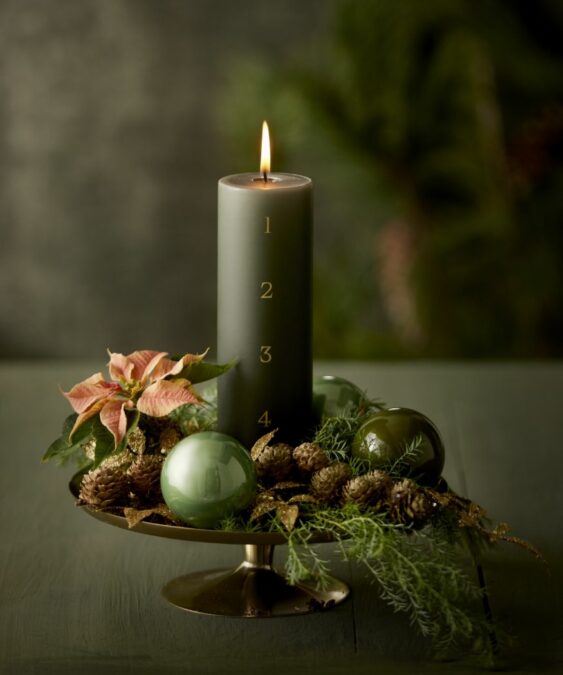Egy modern adventi koszorú alternatíva egyetlen sötétzöld gyertyával, zöld díszekkel és egy rózsaszín mini mikulásvirággal. 