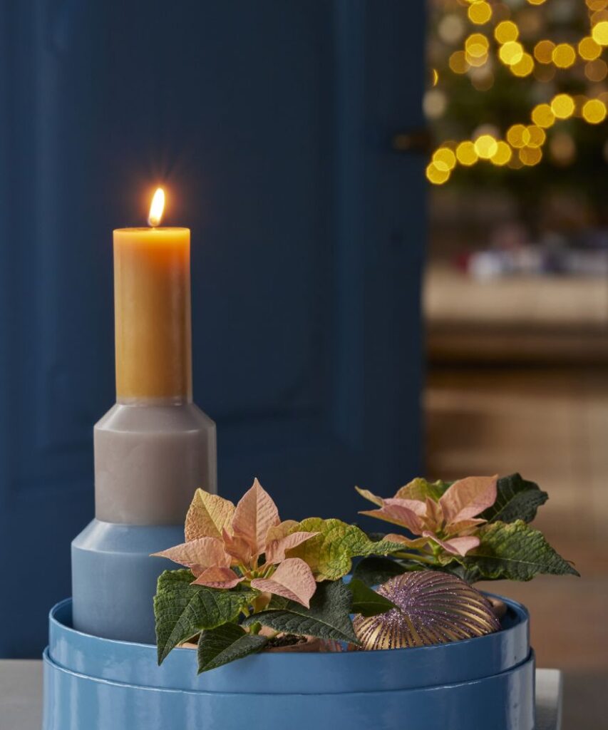 Egy égő oszlopgyertya, amit rózsaszín mikulásvirág és egy nagy díszgömb vesz körül, egy modern kék tálcán elrendezve. A háttérben lévő fények meghitt, ünnepi hangulatot teremtenek. 