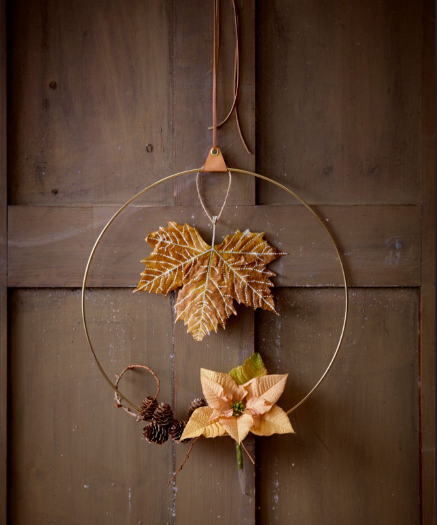 2020-poinsettia-04 Goldener Ring, der mit Lederband an einer rustikalen Tür aufgehängt und mit einem apricotfarbenen Schnittweihnachtsstern im Reagenzglas, einem bemalten Ahornblatt und Lärchenzapfen dekoriert ist