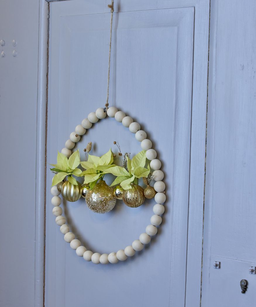 2024_Poinsettia_01110_DIY_Bead_Wreath_Gold_Accents_08 Una ghirlanda di perline di legno finita è appesa a una porta, decorata con palline dorate e Stelle di Natale giallo crema.