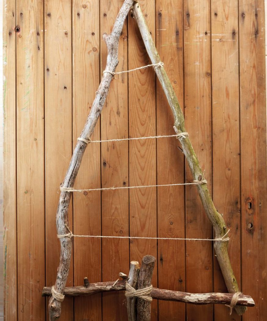 2020-poinsettia-02140-diy-wall-hanging-tree-branches-an-poinsettia-03 Un albero di Natale rustico fatto di rami di legno disposti a triangolo e legati insieme con uno spago grosso. È appoggiato a un pannello di legno.