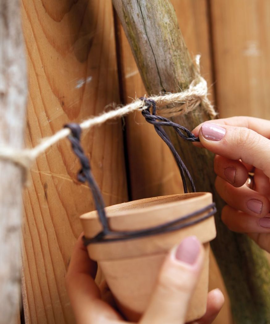 2020-poinsettia-02140-diy-wall-hanging-tree-branches-an-poinsettia-04 Primo piano delle mani che attaccano un piccolo vaso di terracotta allo spago del telaio di rami con un filo di ferro.