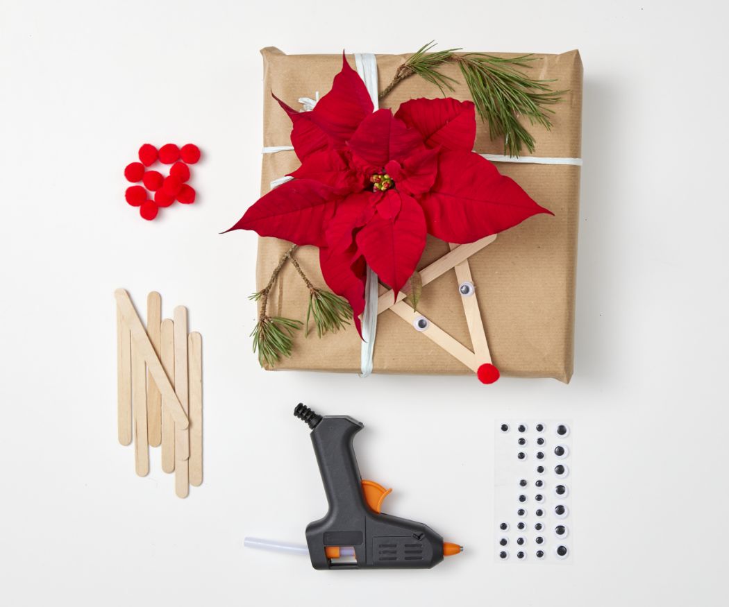 2023-poinsettia-03170-diy-reindeer-gift-trim-01 Materiale per realizzare decorazioni regalo di renne e Stelle di Natale, compresi bastoncini di legno, pistola per colla a caldo e occhietti traballanti.