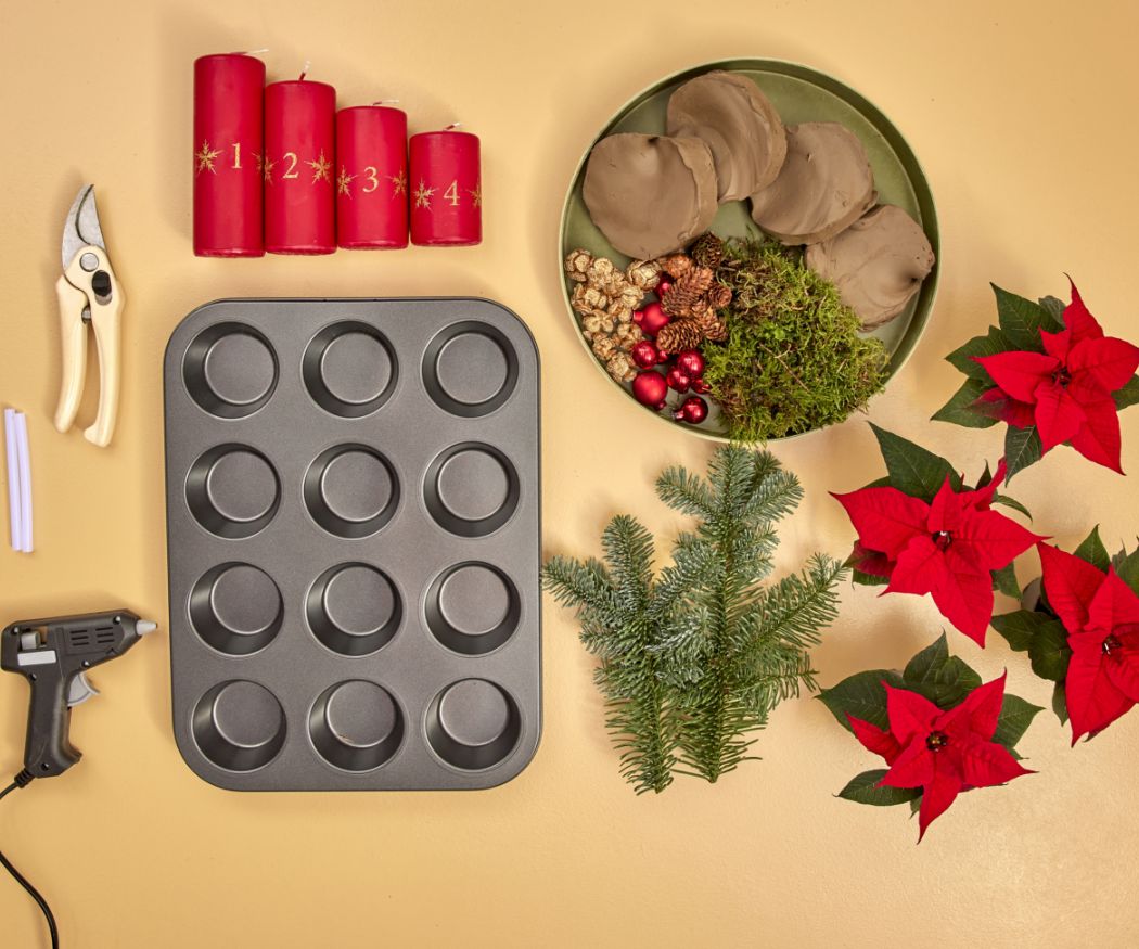 2024-poinsettia-04160-diy-advent-muffin-tin-centrepiece-01 Materiali per una decorazione dell'Avvento fatta in casa in una teglia per muffin, tra cui candele rosse, mini Stelle di Natale, muschio, rametti, argilla e elementi decorativi.