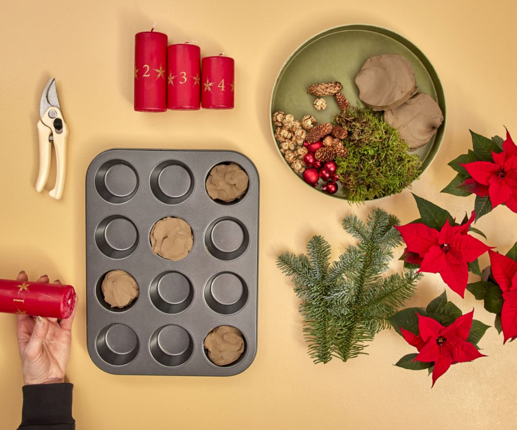 2024-poinsettia-04160-diy-advent-muffin-tin-centrepiece-02 Una candela rossa dell'Avvento viene premuta in uno stampo per muffin con dentro dell'argilla.