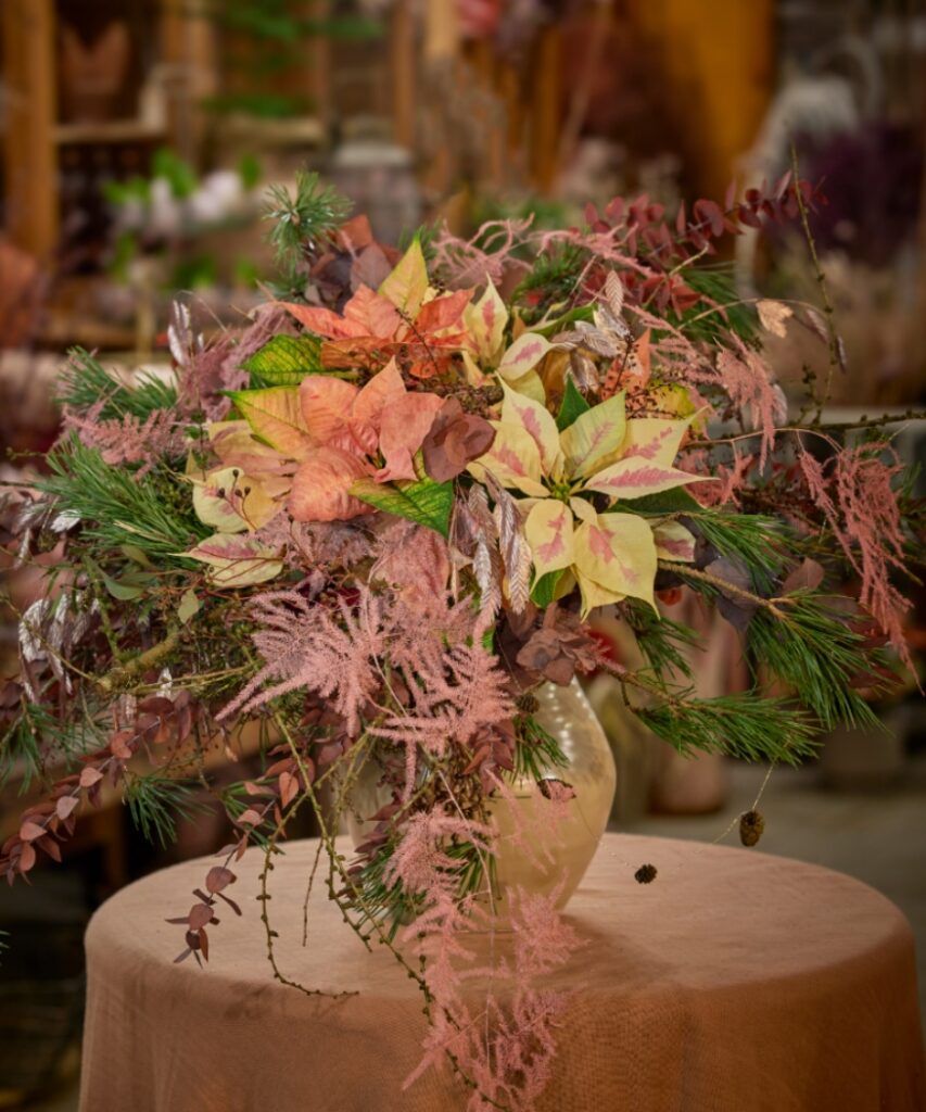 Un grande vaso contenente un'opulenta composizione di Stelle di Natale recise, rami sempreverdi ed elementi floreali secchi è posizionato su un tavolo rotondo coperto da una tovaglia. 