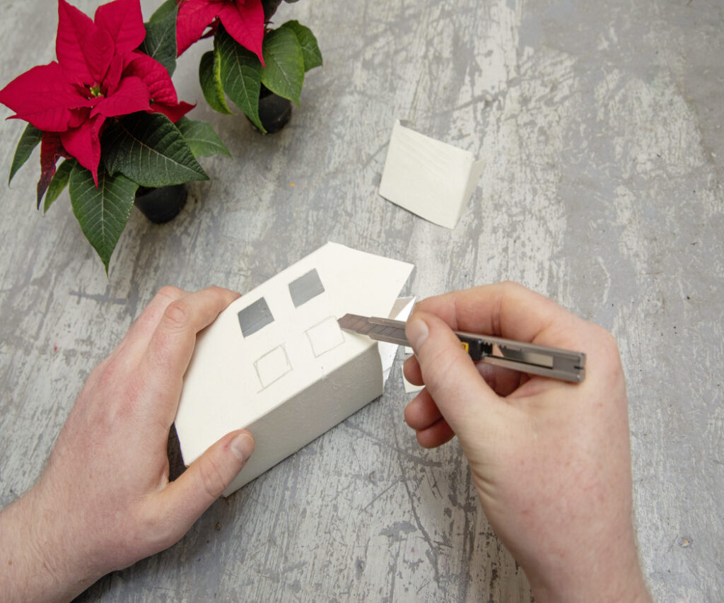 Una persona usa un cutter per ritagliare piccole finestre su un cartone per bevande, nell’ambito di un progetto creativo per realizzare una casetta fai-da-te. 