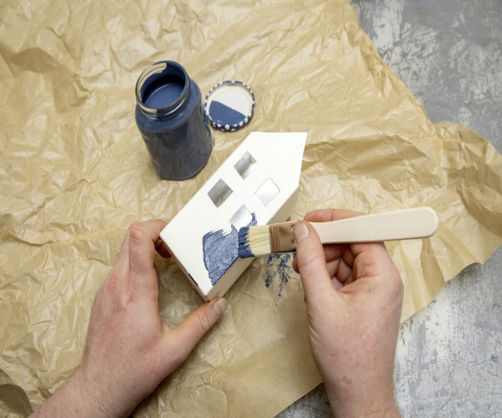 Una casetta di cartone, realizzata con materiali di riciclo, viene dipinta di blu. Passo dopo passo, si dà vita a un villaggio festivo in miniatura. 