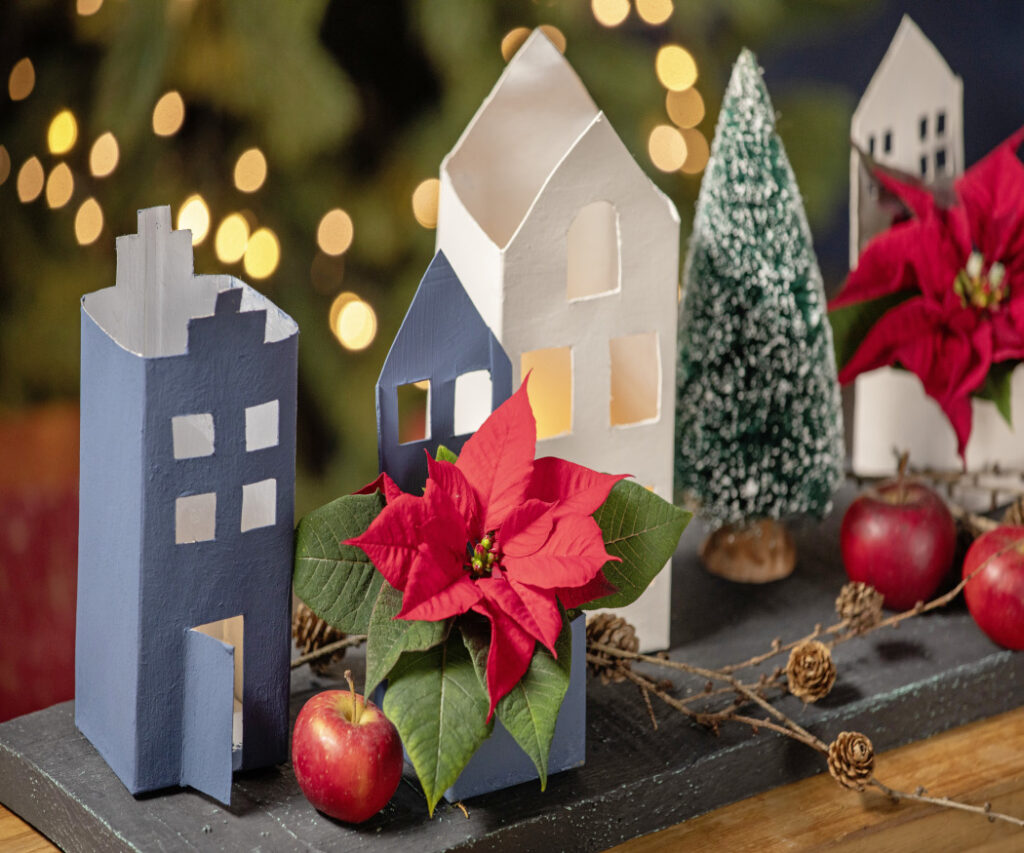 Un primo piano del villaggio realizzato con materiali di riciclo mostra le casette di cartone dipinte, le mini Stelle di Natale e le decorazioni naturali, creando un'atmosfera davvero natalizia. 