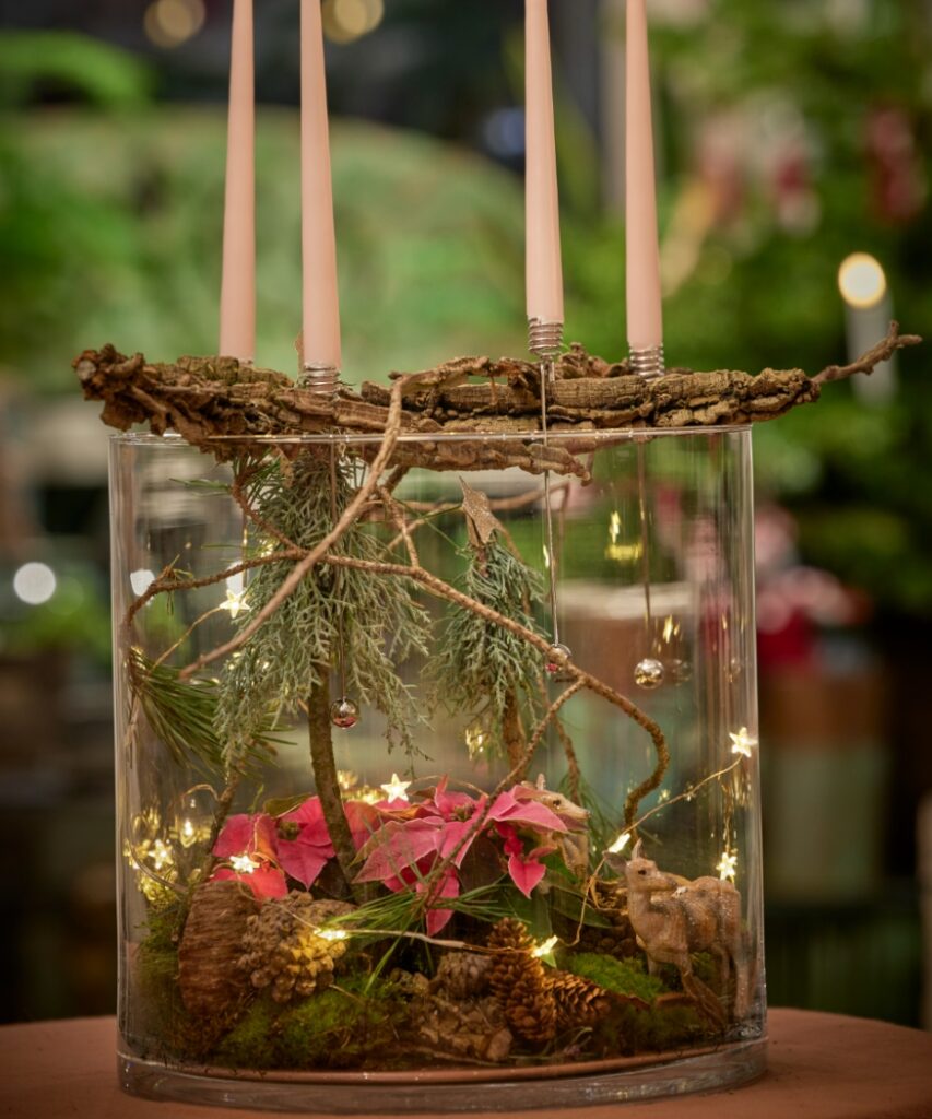 Een adventsdecoratie met een winters tafereel in een glazen schaal, met mini-kerststerren, mos, dennenappels, een hertenfiguurtje en vier witte kaarsen.