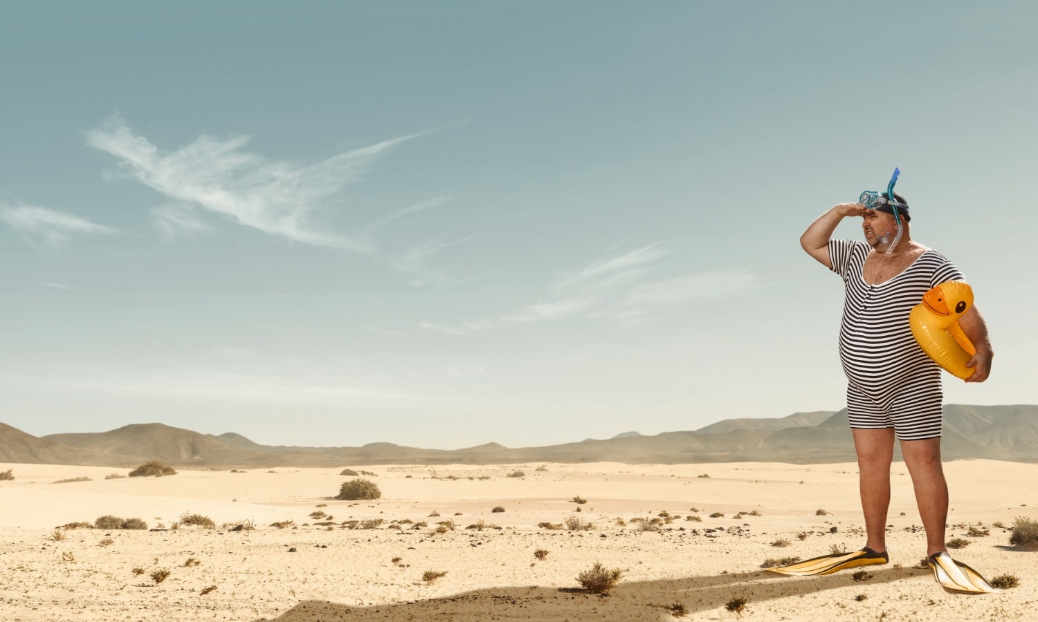 Mann mit Gummiente, Schnorchel und Schwimmfüßen in der Wüste.